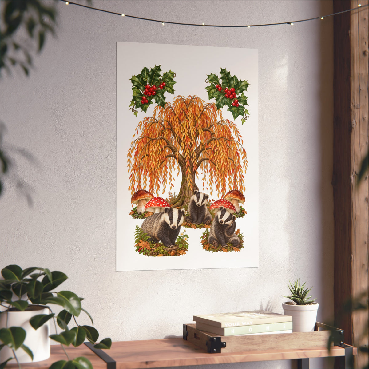 A vertical wall art print featuring an illustration of badgers, a weeping willow tree, and red berries, displayed on a textured white wall above a wooden console table with plants and books.