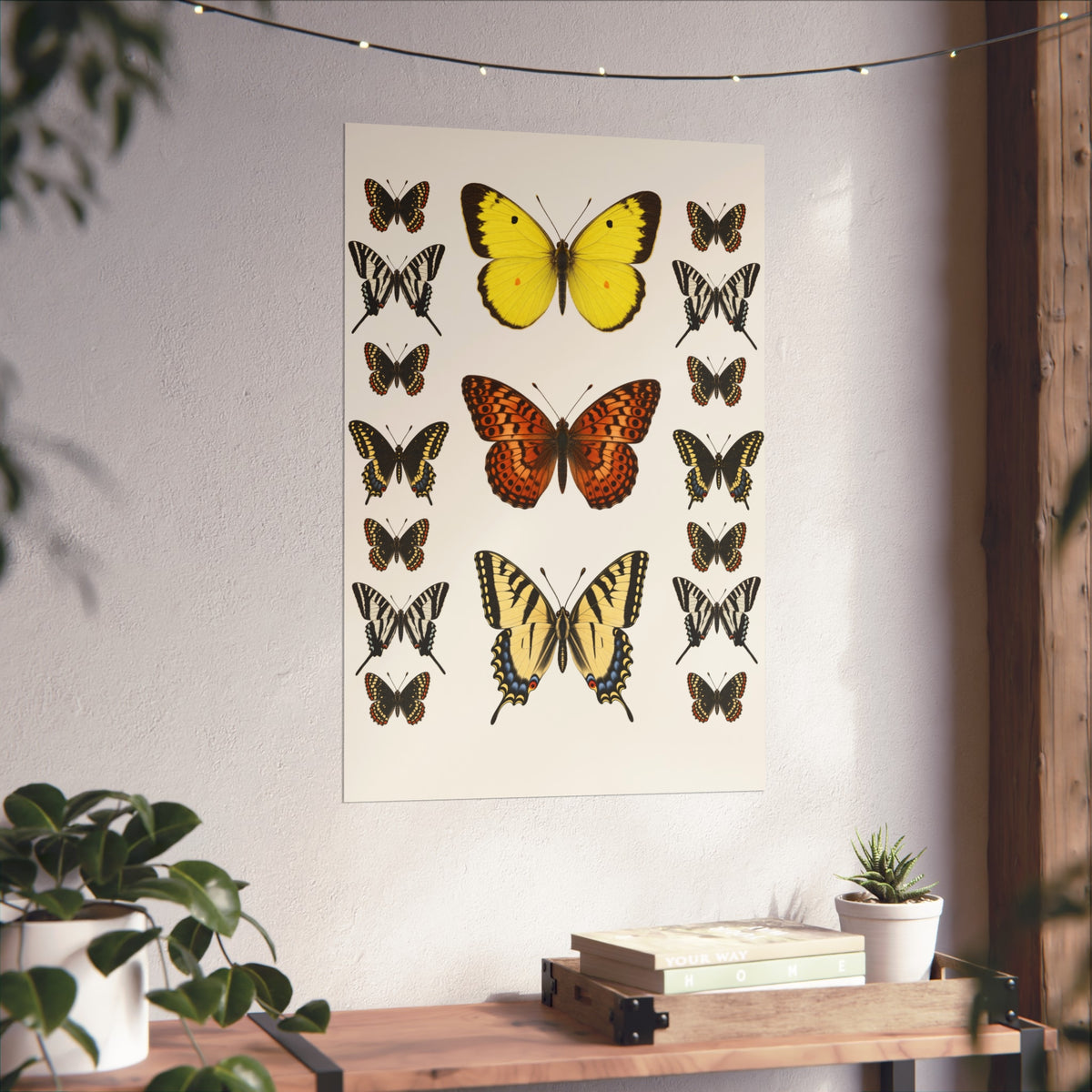 A vertical wall art print featuring vintage-style butterfly illustrations in yellow, orange, black, and cream tones, displayed on a textured white wall above a wooden console table with books and a small plant.