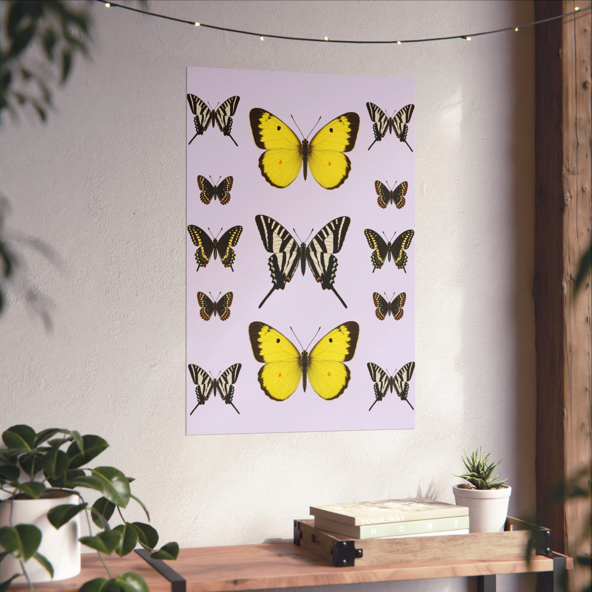 A vertical wall art print featuring yellow and black butterfly illustrations on a pale lavender background, displayed on a textured off-white wall above a wooden console table with books and small plants.