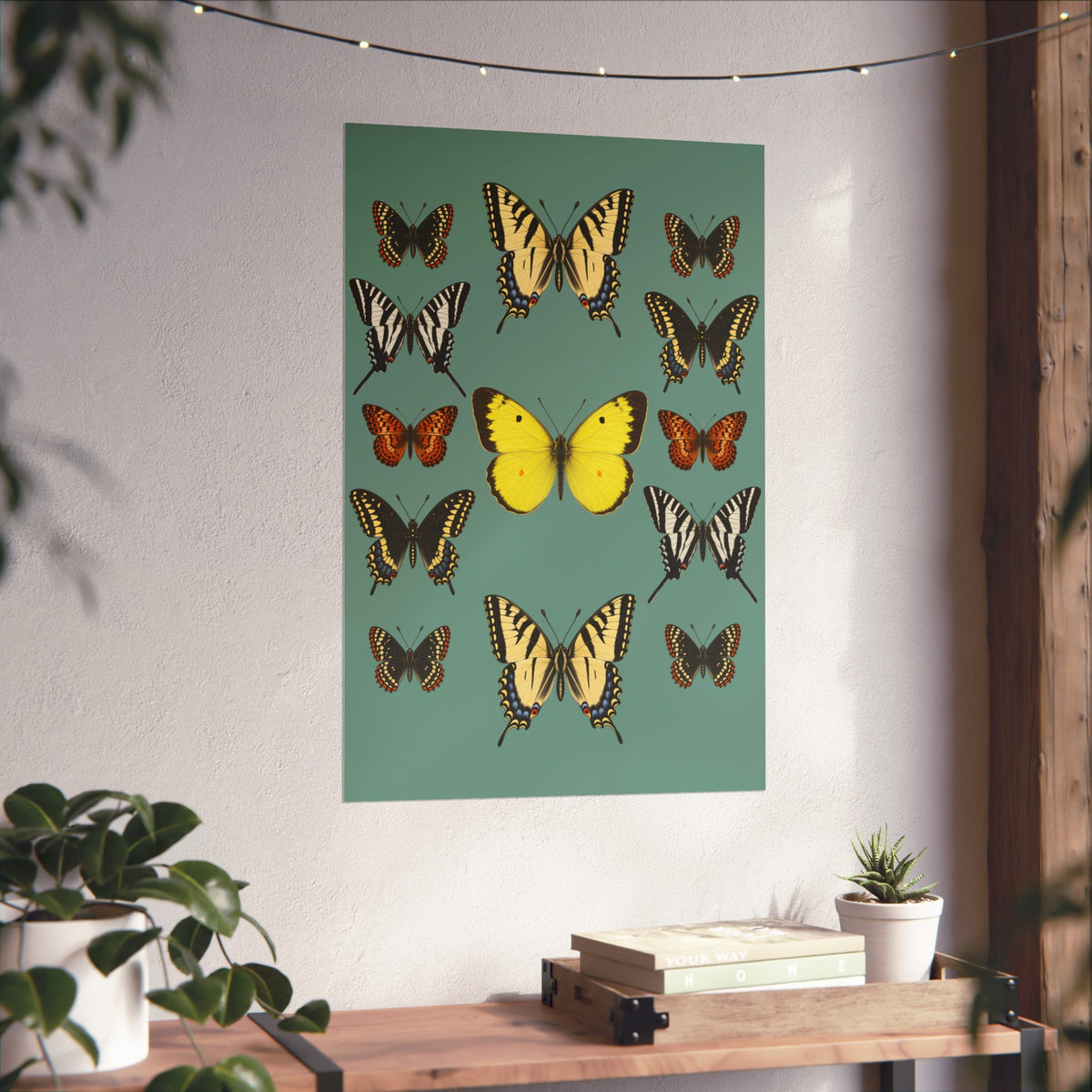 A vertical wall art print featuring multiple illustrated butterflies on a muted teal background, displayed on a white wall above a wooden console table with books and a small plant.