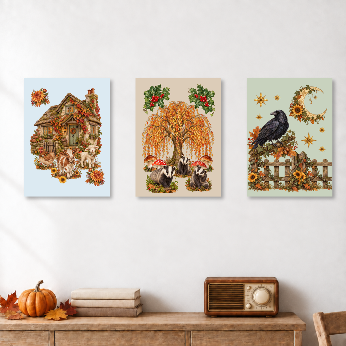 Three vertical autumn-themed art prints displayed side-by-side on a white wall above a wooden console table decorated with a pumpkin, folded blankets, and a vintage radio.