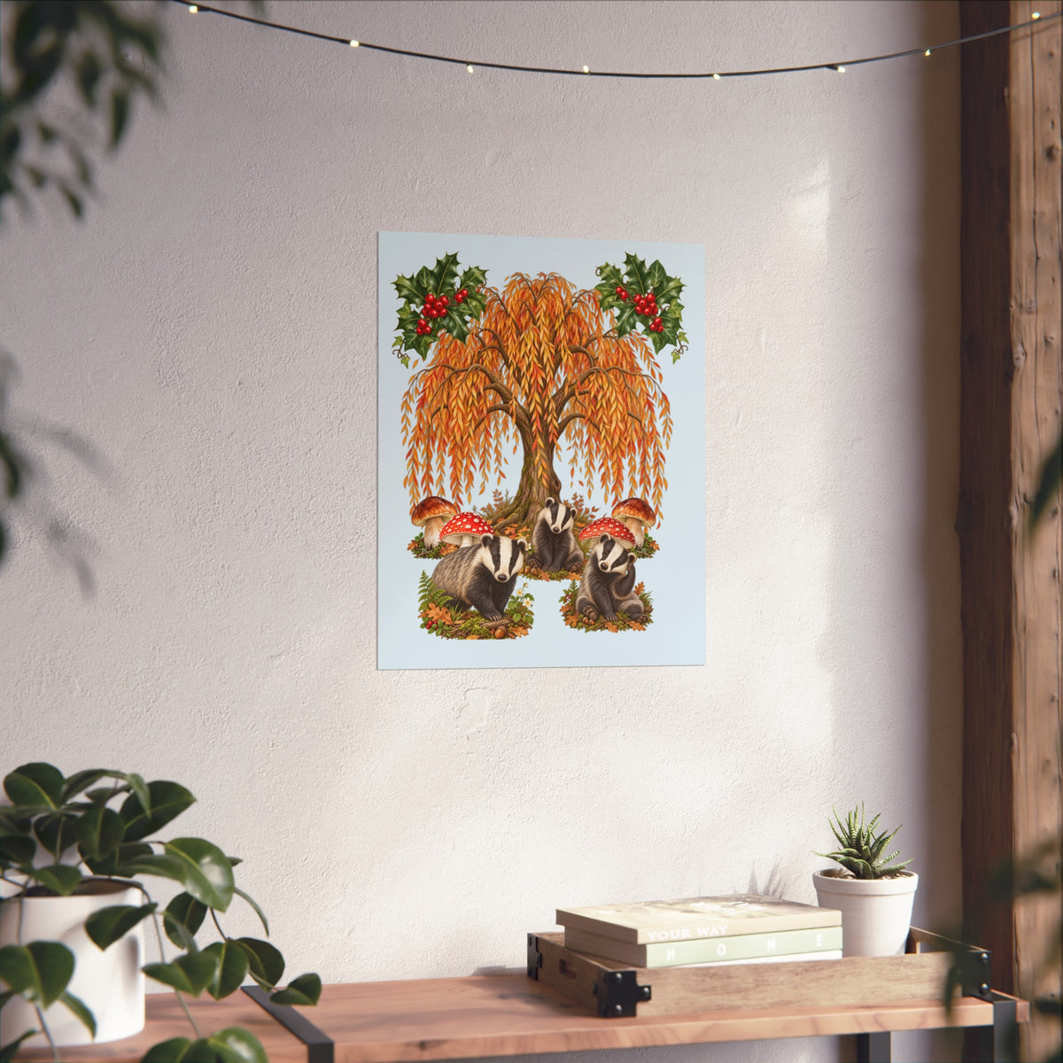 A vertical art print featuring a whimsical autumn tree with orange-yellow leaves, red berries, and badgers among mushrooms, displayed on a textured white wall above a wooden shelf with plants.