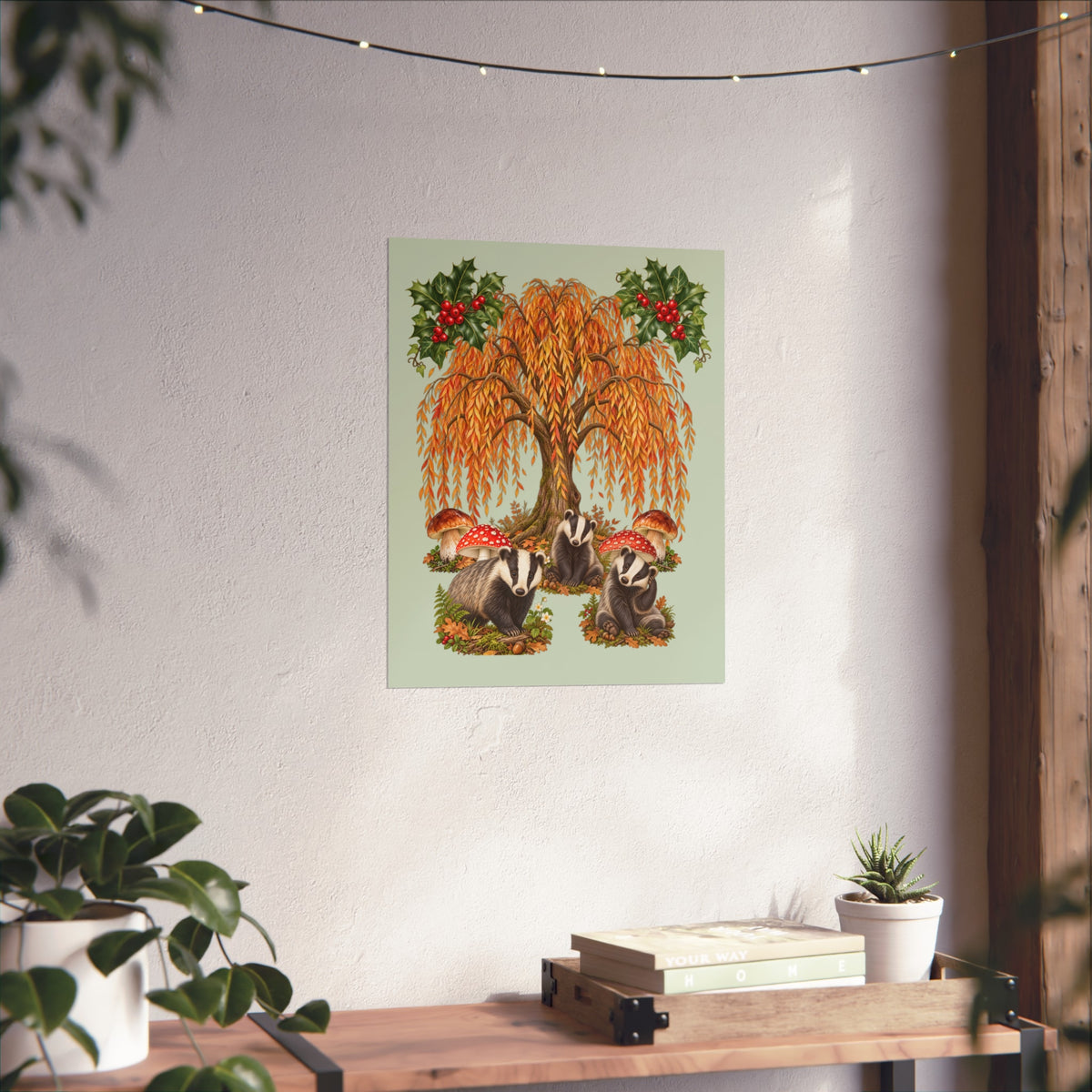 A vertical art print featuring a weeping tree with orange-yellow leaves, red berries, and badgers at its base, displayed on a textured white wall above a wooden shelf with books and plants.