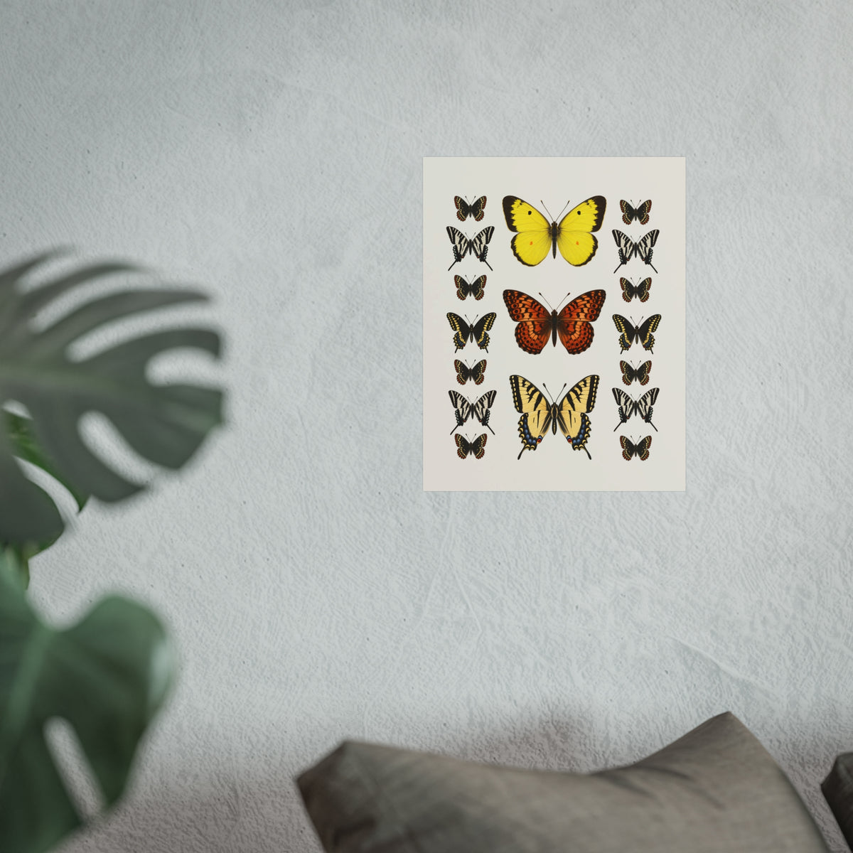 A vertical art print featuring various illustrated butterflies on an off-white background, displayed on a textured light gray wall with a blurred green plant leaf on the left and a gray pillow at the bottom.