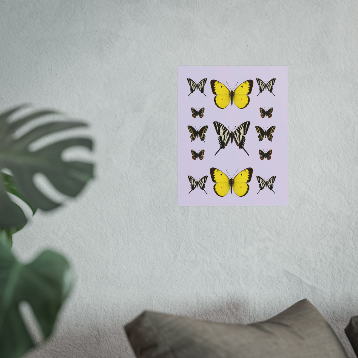 A rectangular wall art print featuring multiple butterfly illustrations, including two prominent yellow butterflies and several black-and-white striped species, displayed on a textured light gray wall above a sofa with a blurred plant in the foreground.