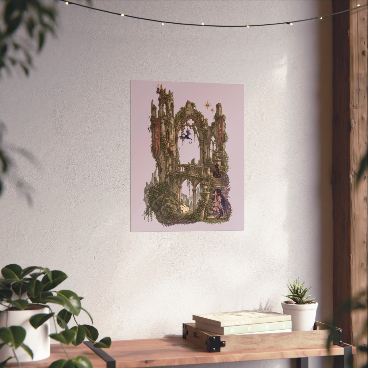 A vertical art print featuring a whimsical, fantasy-style illustration of a mossy stone tower and archway under a pale pink sky, displayed on a textured white wall above a wooden console table with plants