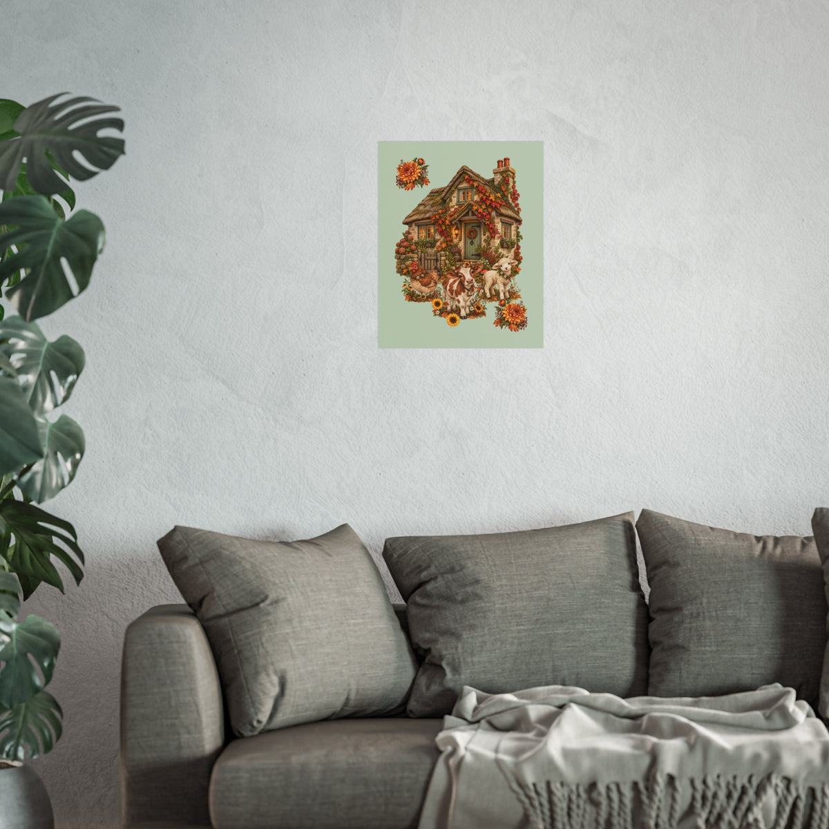 A framed or unframed wall art print depicting a small cottage surrounded by autumn leaves and animals, displayed on a textured light gray wall above a gray sofa with pillows and a throw blanket, with a large green plant on the left.