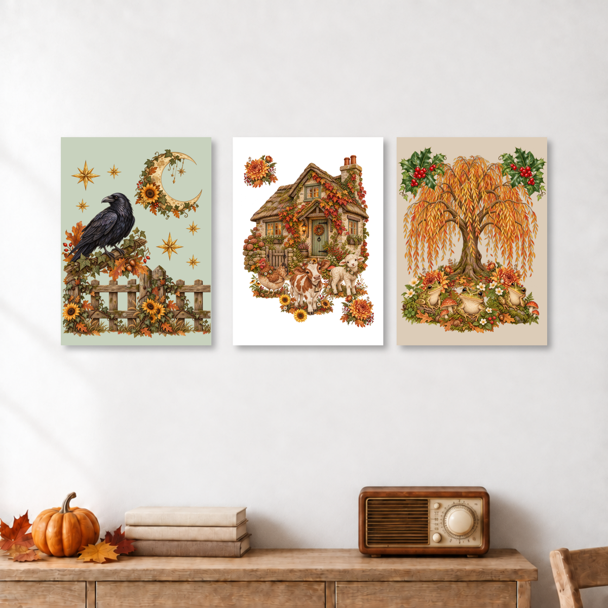 Three vertical art prints with autumn-themed illustrations are displayed on a white wall above a wooden console table. The left print shows a black raven, sunflowers, and a crescent moon on a pale green background.