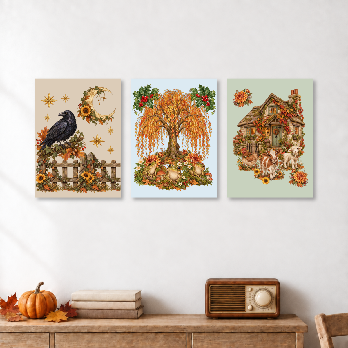 Three vertical art prints with autumn and cottage themes are displayed on a white wall above a wooden console table with a pumpkin, books, and a vintage radio.