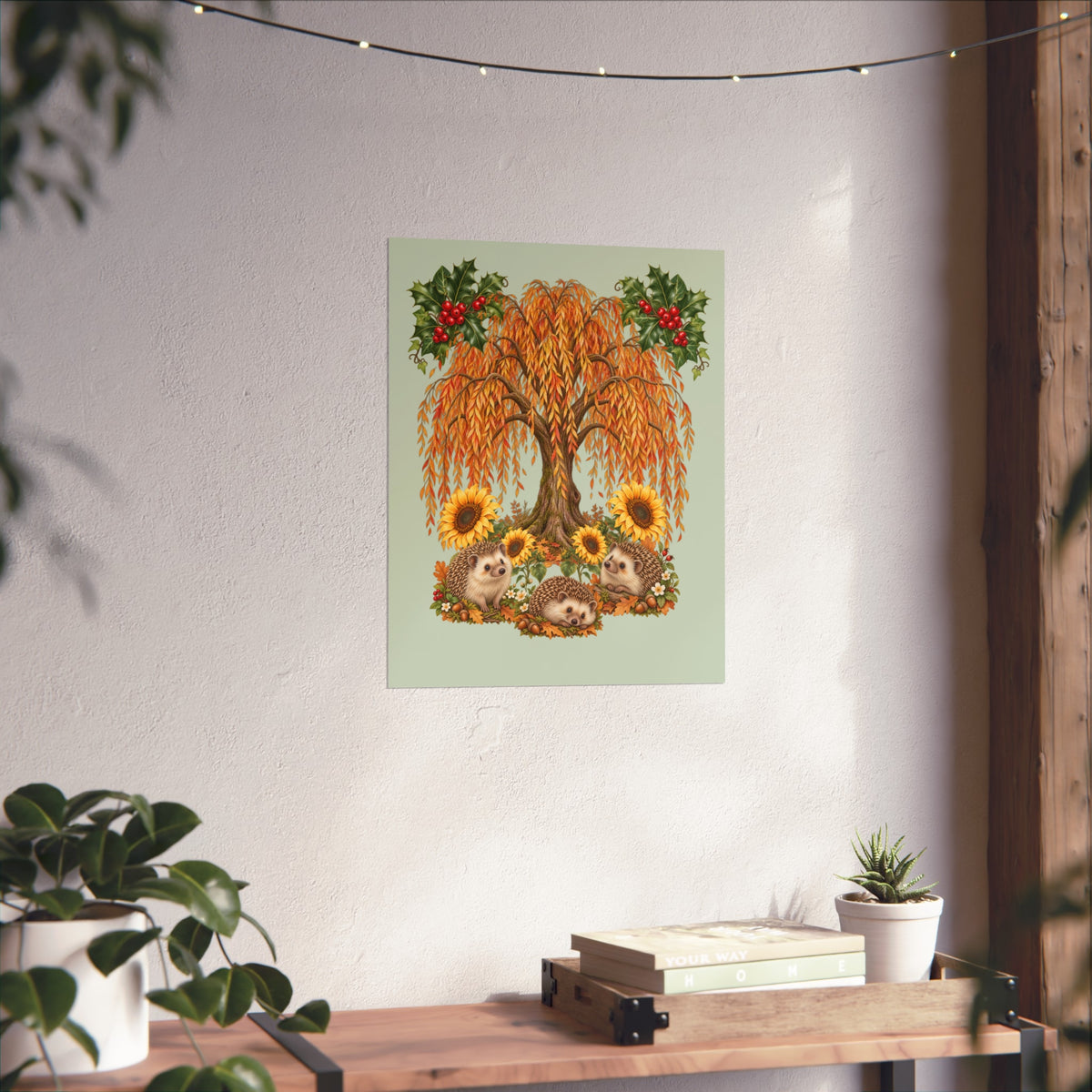 A vertical art print featuring a weeping willow tree with sunflowers and hedgehogs, displayed on a textured white wall in a styled room setting with a wooden shelf, plants, and string lights.