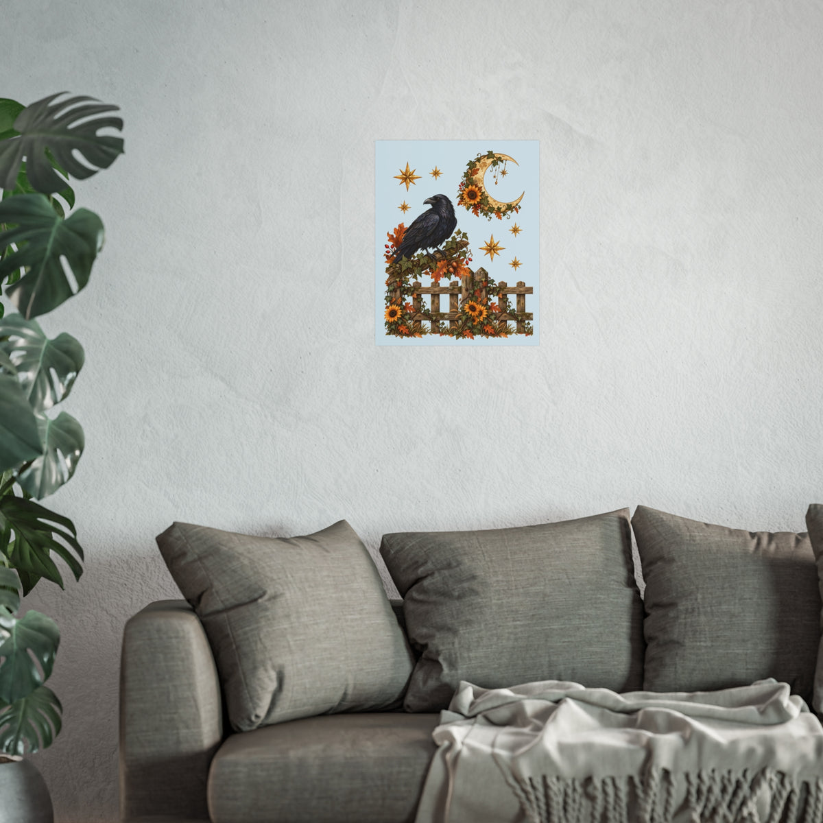 A small square wall art print featuring a black raven perched on a fence with autumn leaves, stars, and a crescent moon on a pale blue background, displayed above a gray sofa in a living room with a plant at left.