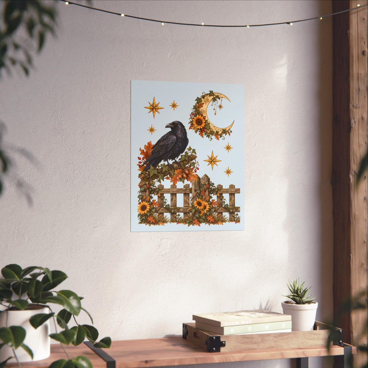 A vertical art print featuring a black raven perched on a wooden fence with sunflowers, autumn leaves, a crescent moon, and stars, displayed on a textured white wall above a wooden console table with plants.