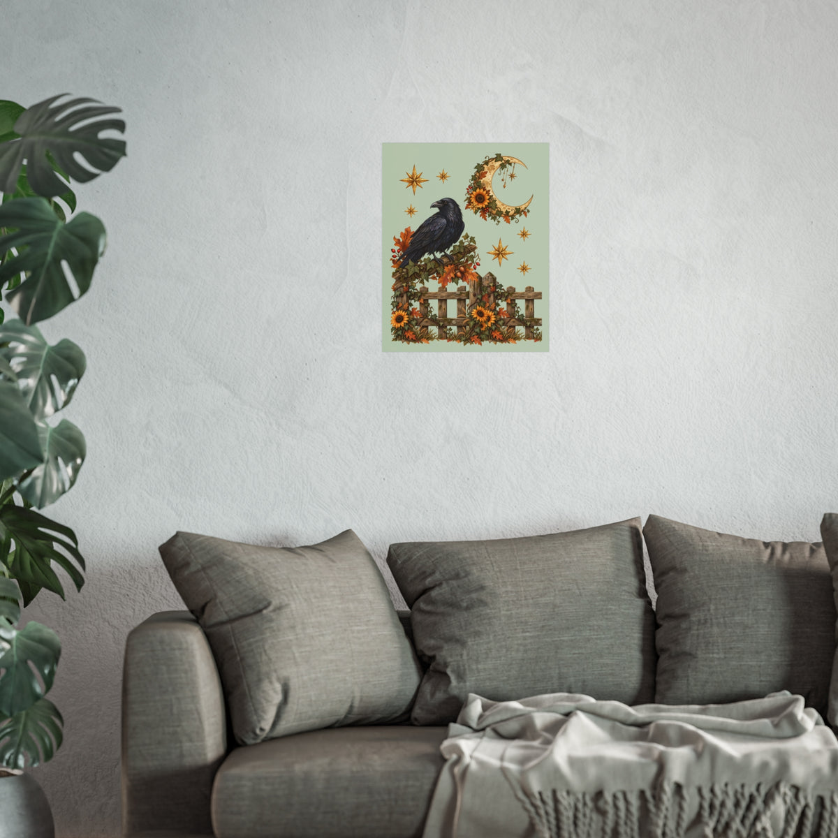 A framed wall art print featuring a black raven perched on a fence with autumn flowers, a crescent moon, and stars, displayed above a gray sofa in a living room setting with a plant on the left.
